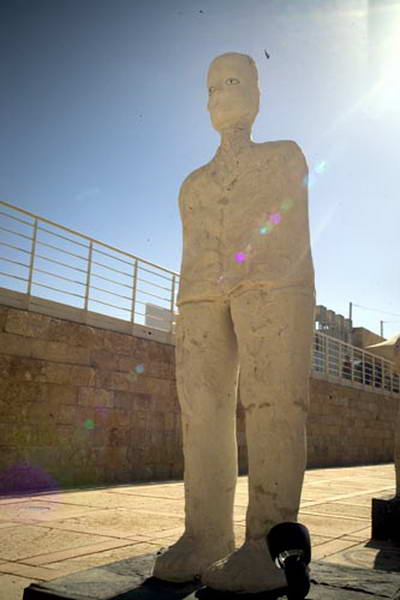 Full view of a tall pale standing figure outdoors, on a dark plinth beside a metal railing.