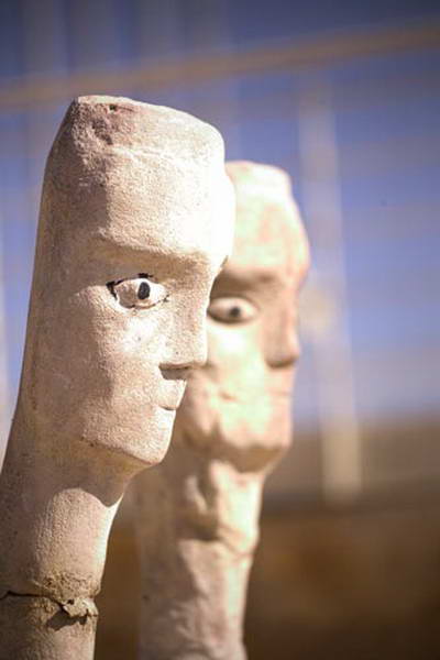 Side view of two elongated sculpted heads, one in the foreground and one behind.