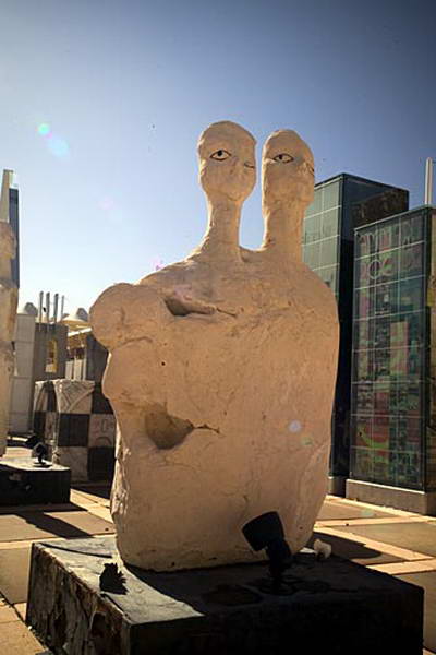 A two-headed sculpture on a dark outdoor plinth, with glass buildings behind it.