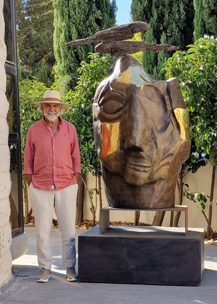 A large bronze head with two birds above it, outdoors among cypresses.