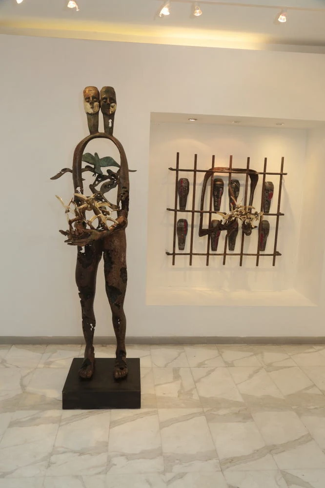 A standing bronze twin figure with birds, and a grille of masks on the wall behind.