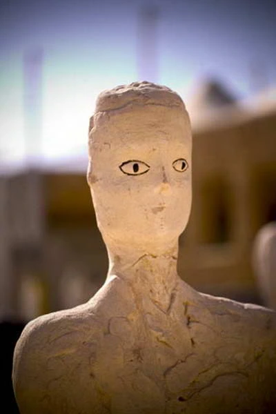 Front view of a pale sculpted face and upper torso with eyes outlined in black.