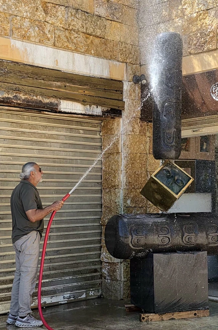 A man at the left sprays water over the tall basalt-and-bronze sculpture outside the gallery.