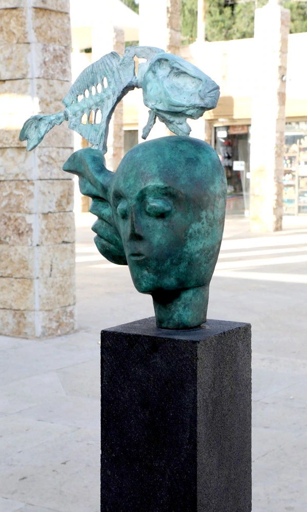 A sculpted head topped by a fish, outdoors; first angle.