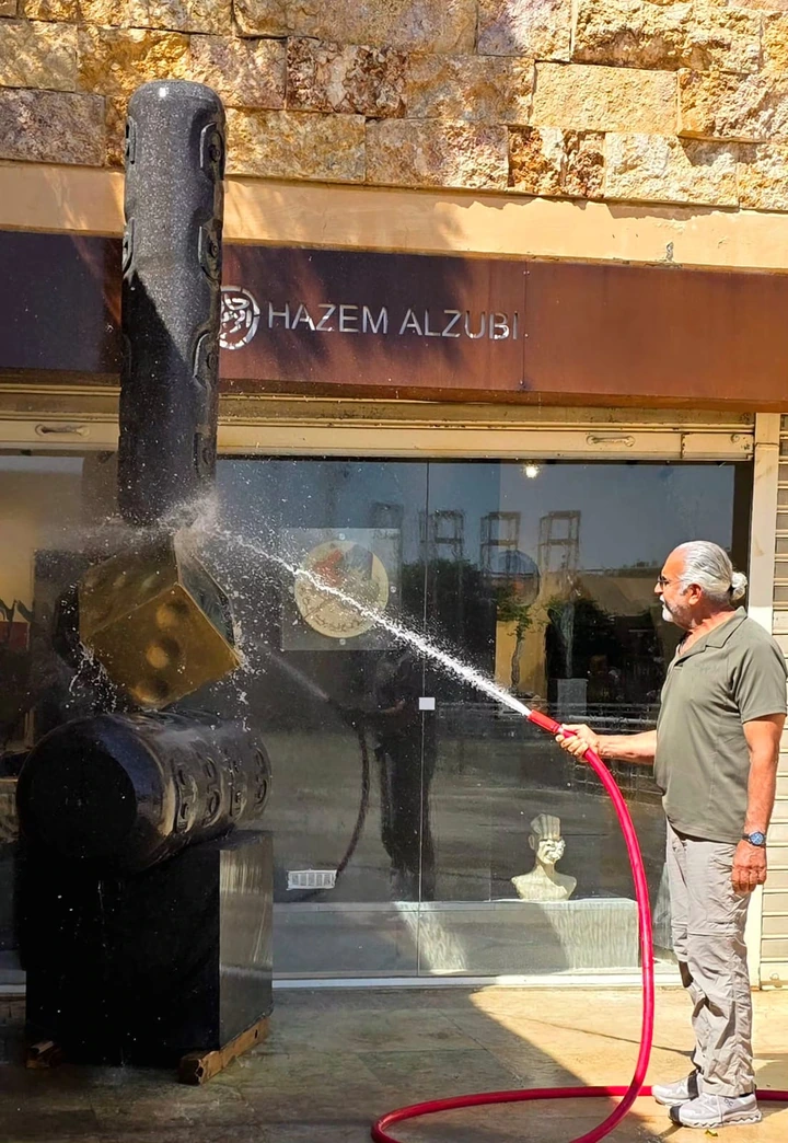 A man at the right washes the sculpture with a red hose, beneath the gallery sign.