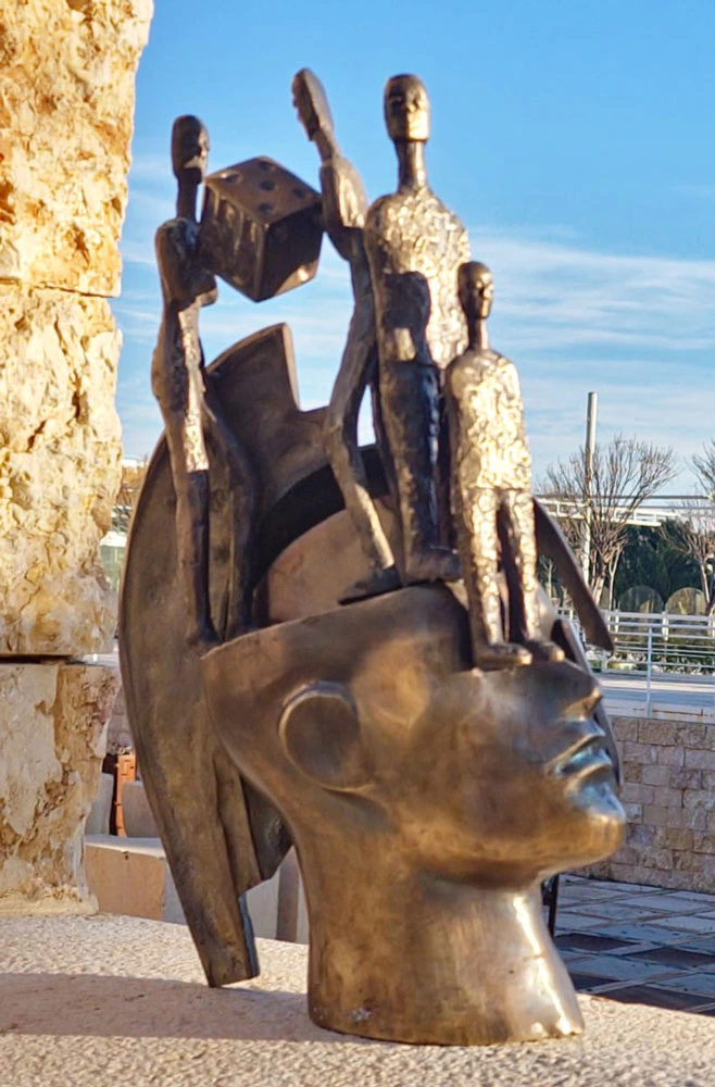 A large head with standing figures and a die, photographed outdoors.