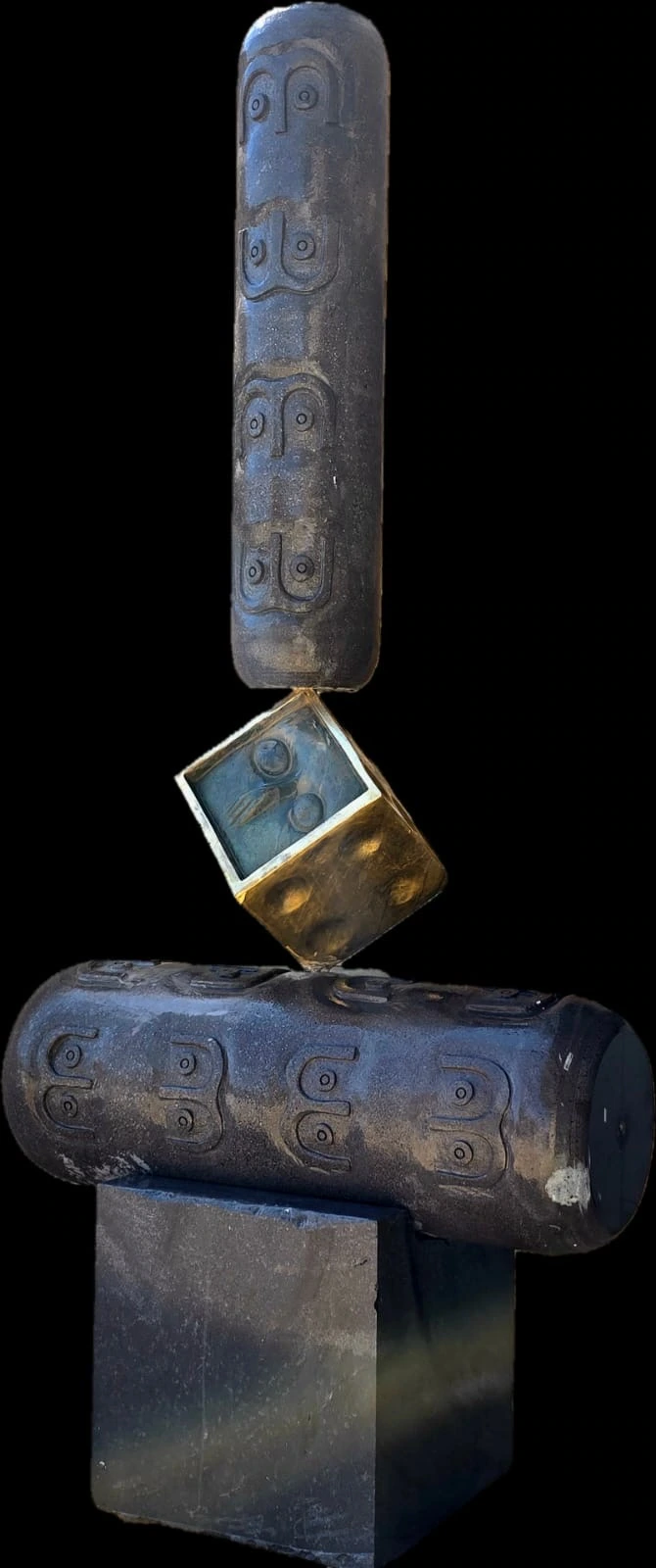 Alternate isolated view of carved basalt cylinders balanced around a golden bronze cube, against black.
