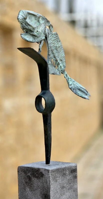 A pierced bronze fish balanced on a thin black steel stalk on a stone plinth.