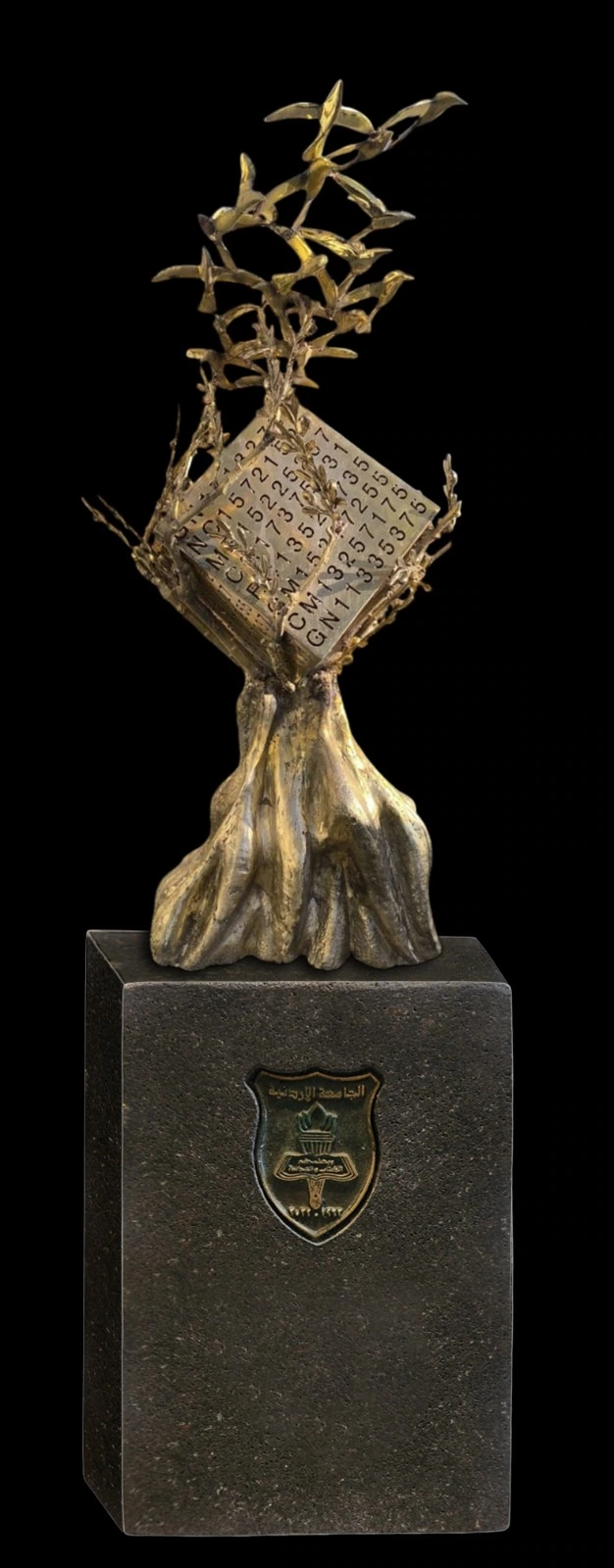 Bronze tree, numbered cube and rising birds on a plinth bearing the University of Jordan crest, against black.
