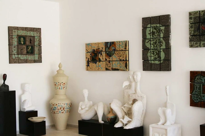 Gallery display of a tall lidded ceramic vessel and pale reclining figures beneath green tiled panels.