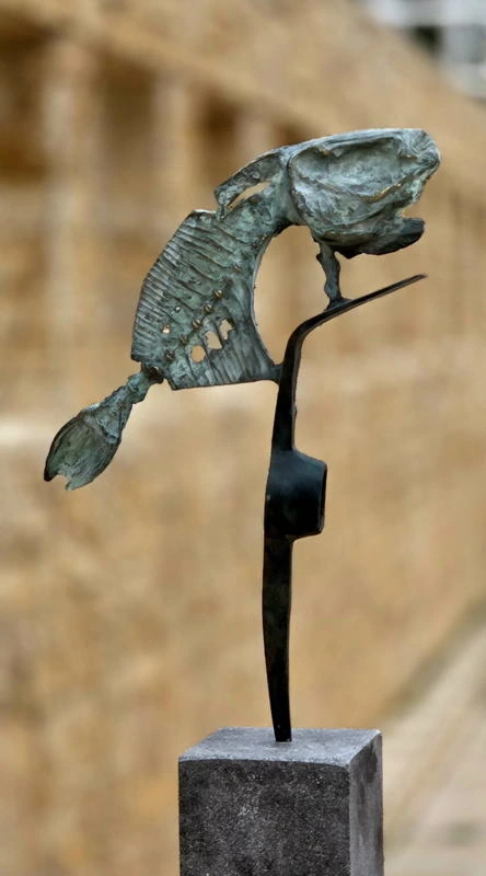 Pierced bronze fish on a thin black support, seen from the opposite side against a blurred stone wall.