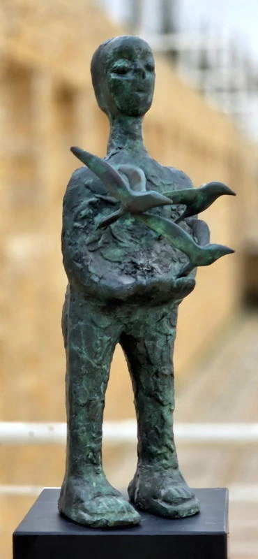 A green-patinated bronze figure standing with both arms curved at the waist, three birds rising out of its cupped hands.
