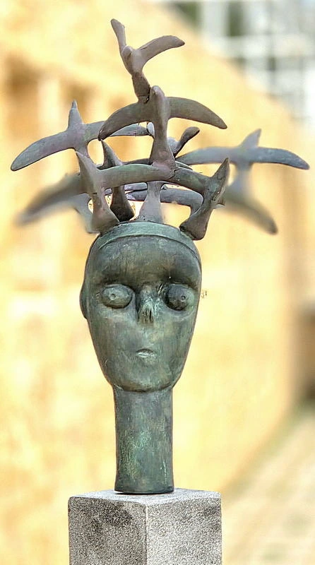 Front view of a green-patinated head crowned by overlapping bronze birds, on a stone plinth outdoors.