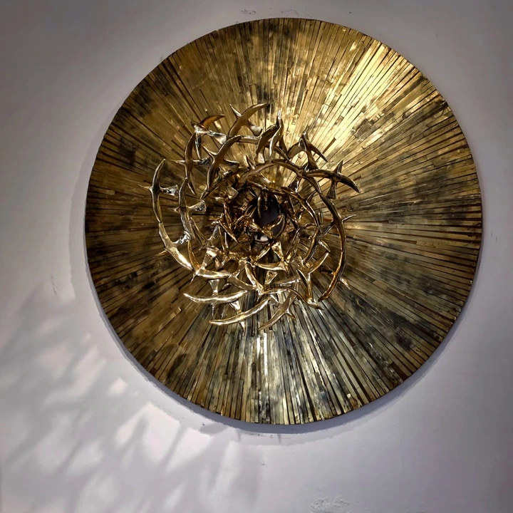 A large circular brass relief, hundreds of narrow rays darkening outward, with a spiral of cast gilded birds converging on a dark opening at the centre.