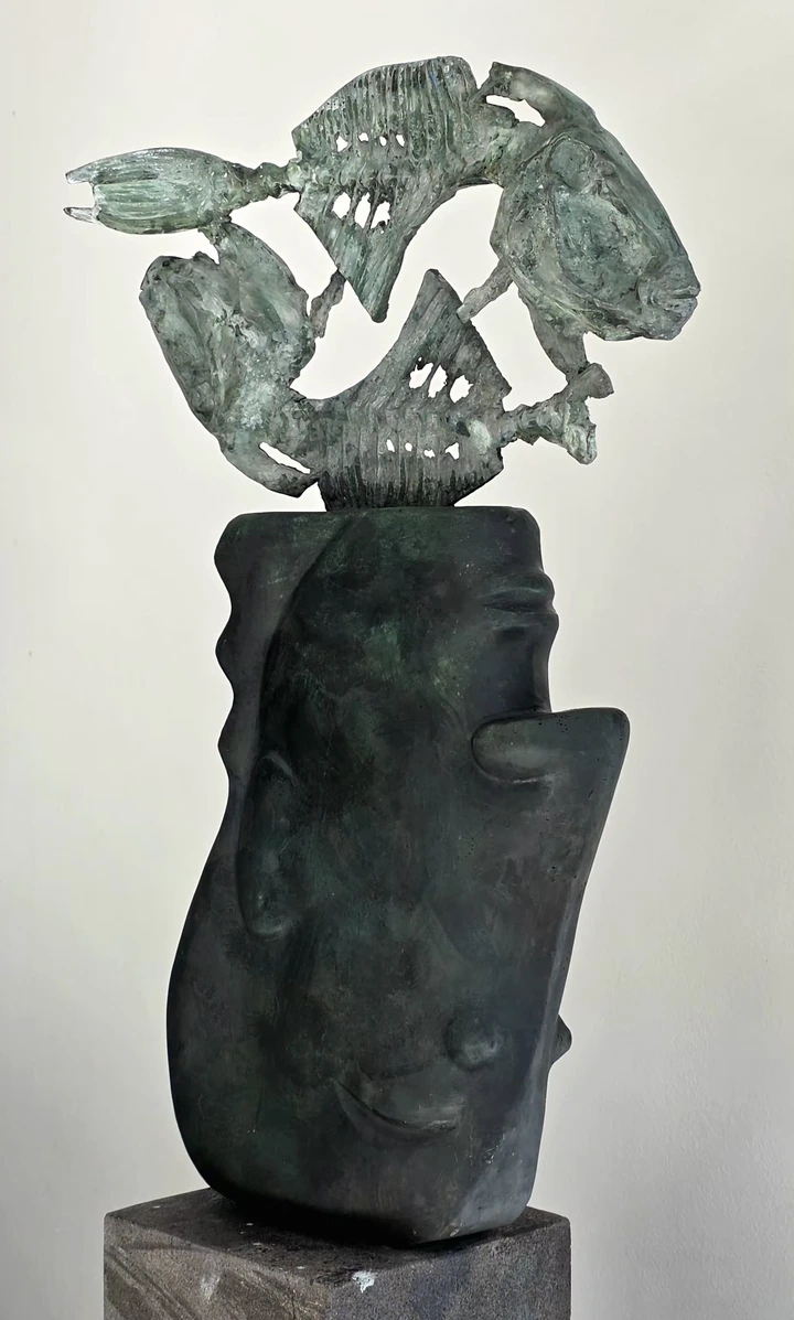 A green-patinated bronze head with pierced fish forms rising above it on a stone plinth.