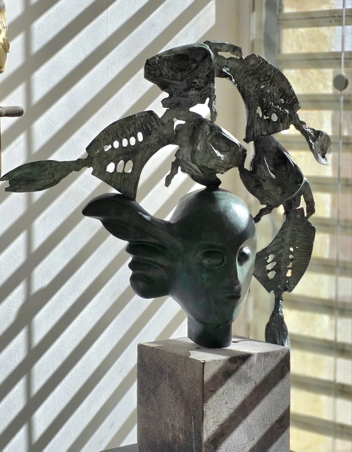 A second bronze head, this one winged, with pierced fish above it, in light thrown through window blinds.