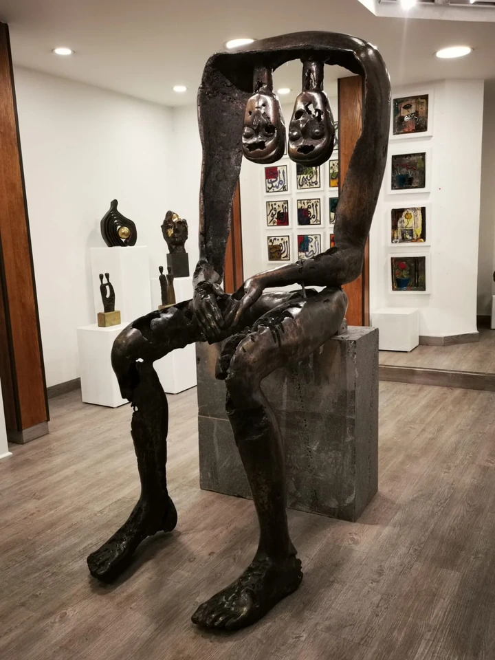 A tall bronze figure seated on a basalt block in a gallery: an open frame for a body, with two pierced heads hanging side by side where the face would be.
