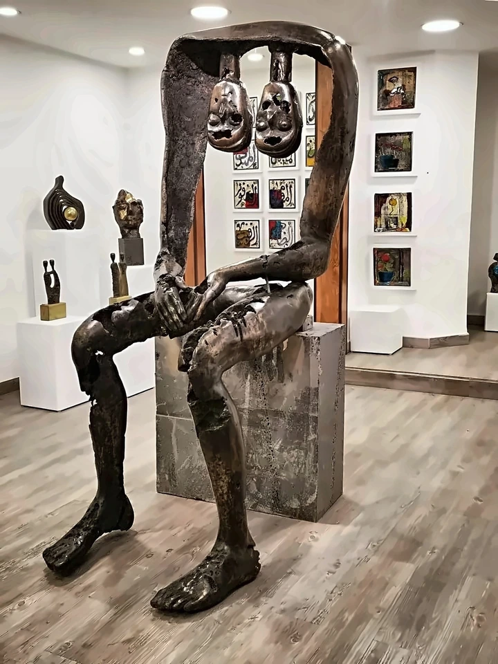 A large seated bronze figure with two heads suspended from a bar above it, on a stone plinth in a gallery.