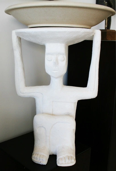 A white plaster figure carrying a wide bowl on its head with both arms raised.