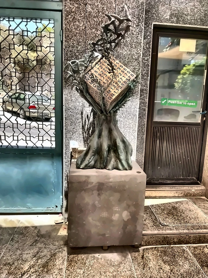 A bronze olive tree on a stone plinth, holding a cube incised with serial numbers, birds rising from the branches, standing inside a doorway of the Central Bank of Jordan.