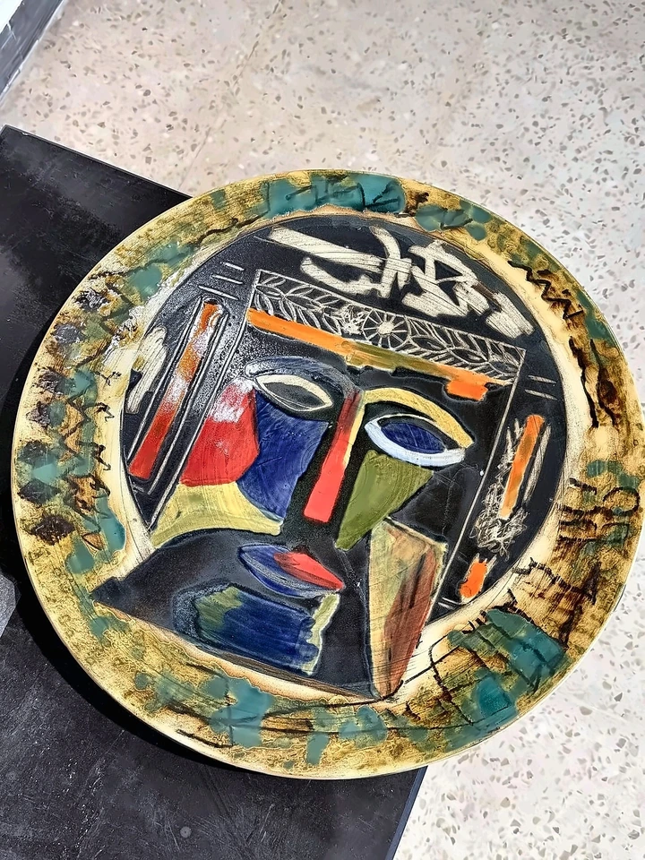 Circular ceramic plate with a stylised face, red nose and blue and yellow glaze.