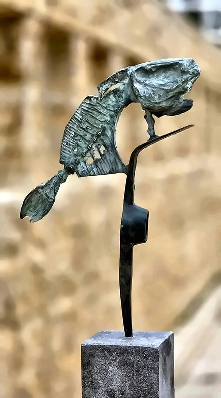 A pierced bronze fish balanced on a thin black steel stalk on a stone plinth.