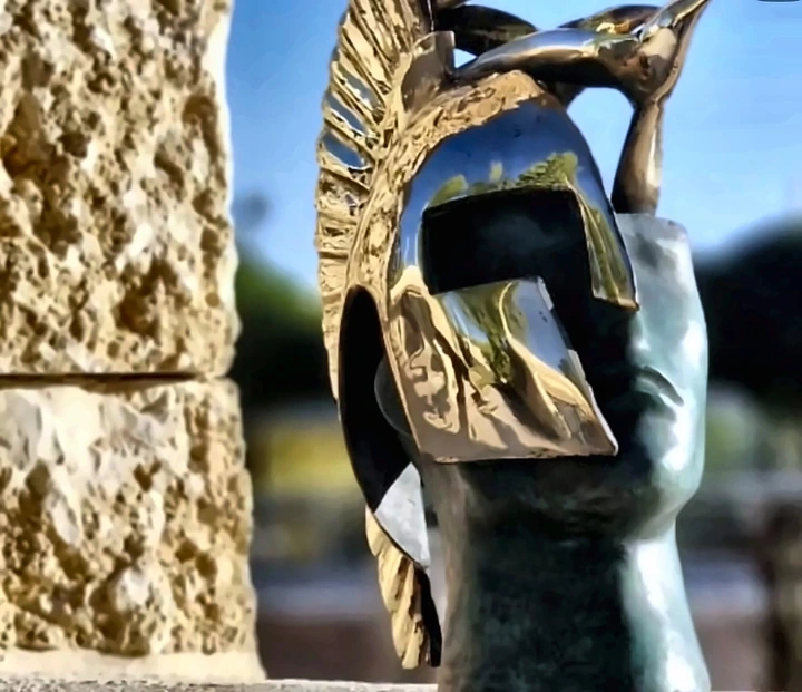 A stone form fitted with a bronze helmet and a bird, against a stone wall.