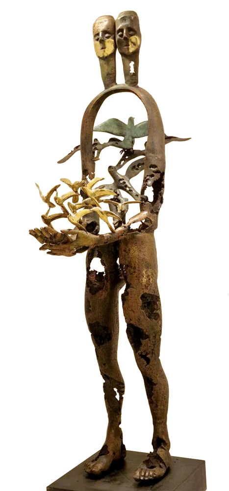 A tall bronze figure with two heads on one neck, its torso an open arch, holding a mass of gilded birds in both hands, the metal pitted and holed throughout.