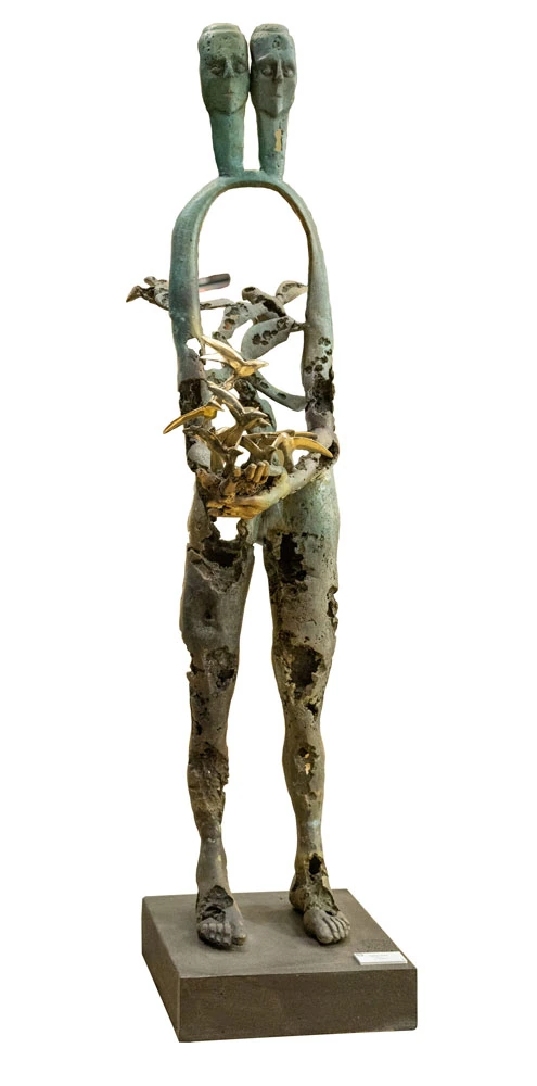 A seated bronze figure in green patina with polished golden feet and shins, two burnished birds resting on the knee.