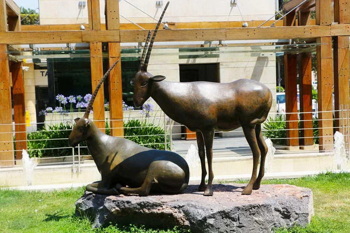 Two bronze Arabian oryx on a block of dark basalt in a landscaped courtyard, one standing with long ribbed horns raised, one lying beside it.