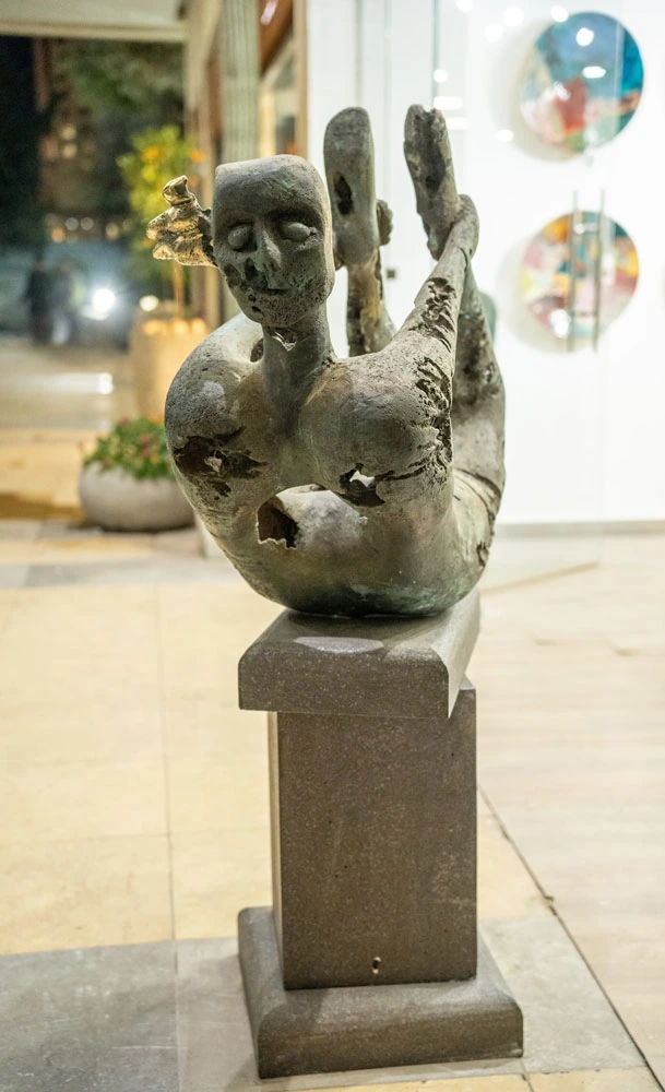 Frontal gallery view of a reclining bronze figure, its perforated torso and small golden birds above a tall plinth.