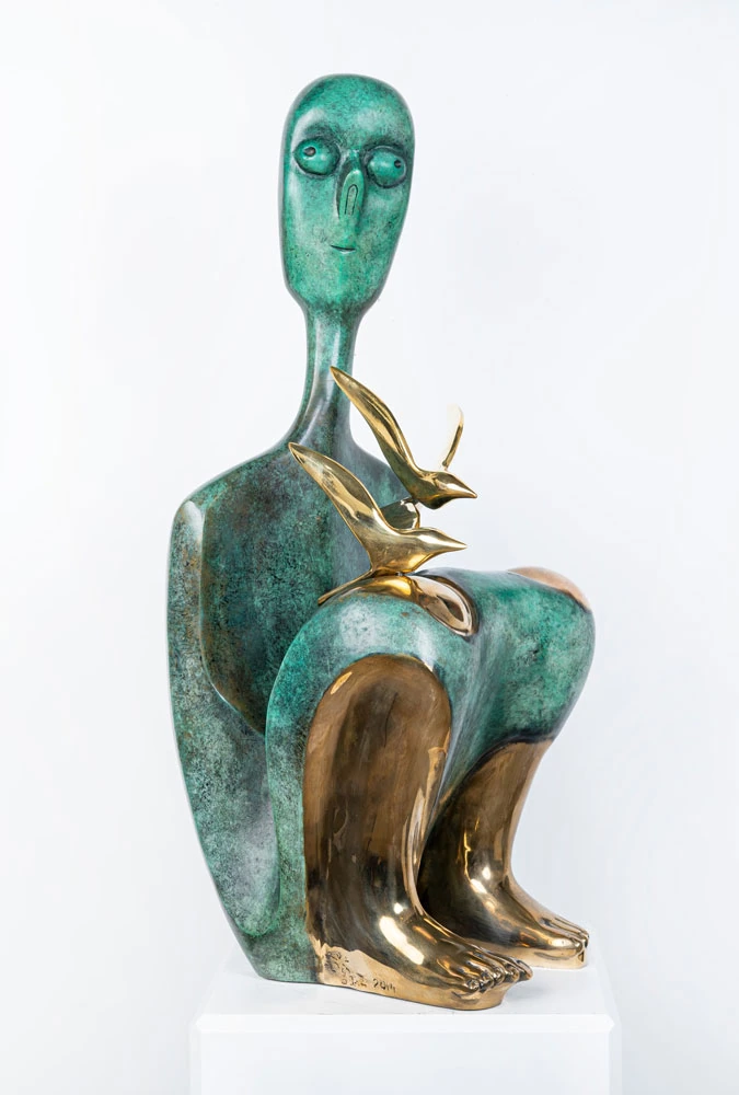 A seated bronze figure in green patina with polished golden feet and shins, two burnished birds resting on the knee.