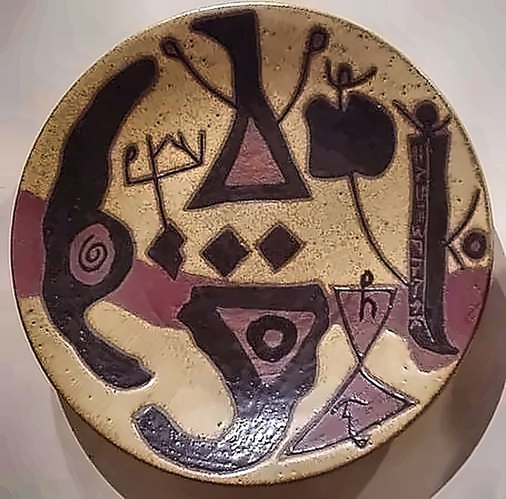 A stoneware plate with heavy black forms, a triangle, a spiral and a column of incised script down one side.