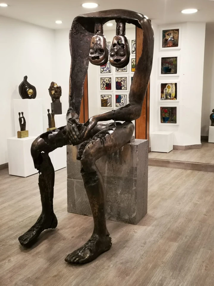 A monumental seated bronze figure in a gallery, its torso an open arch with two small pierced heads hanging inside it, legs at full length, hands folded over one knee.
