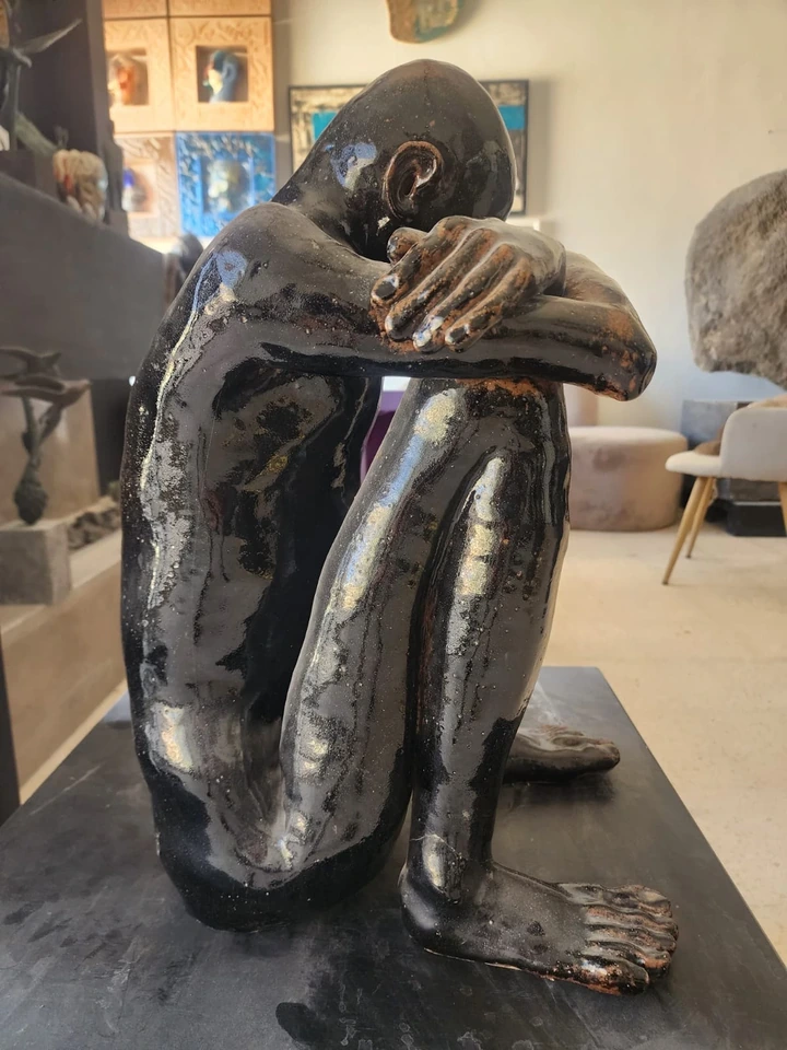A glazed black stoneware figure seated with knees drawn up and head resting on folded arms, seen from the side.