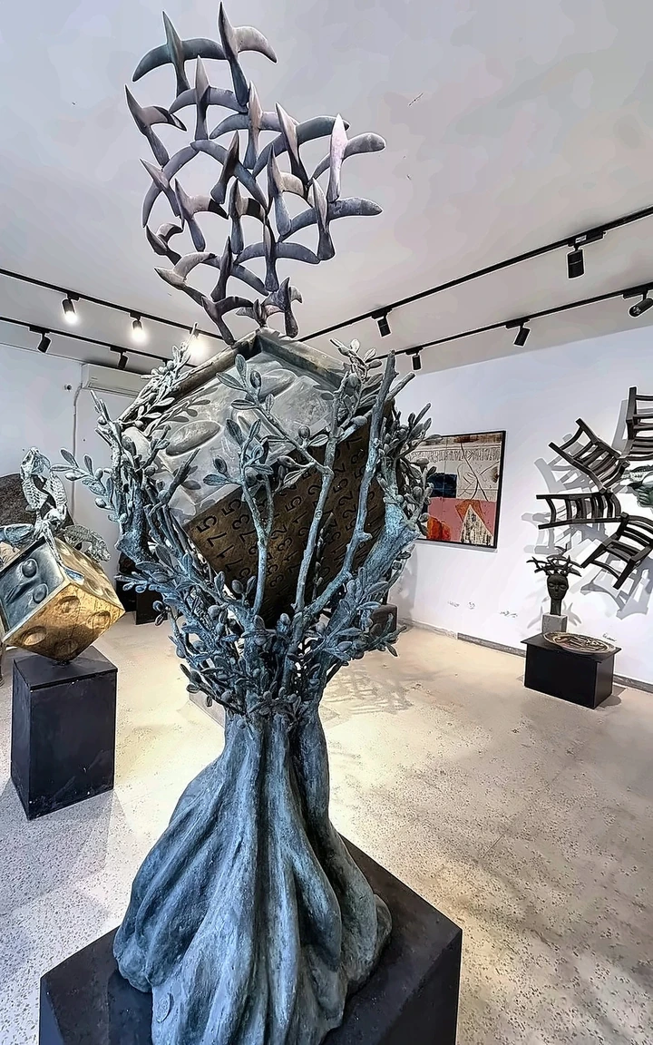 A bronze olive tree with birds lifting from its crown, the gilded die behind it.