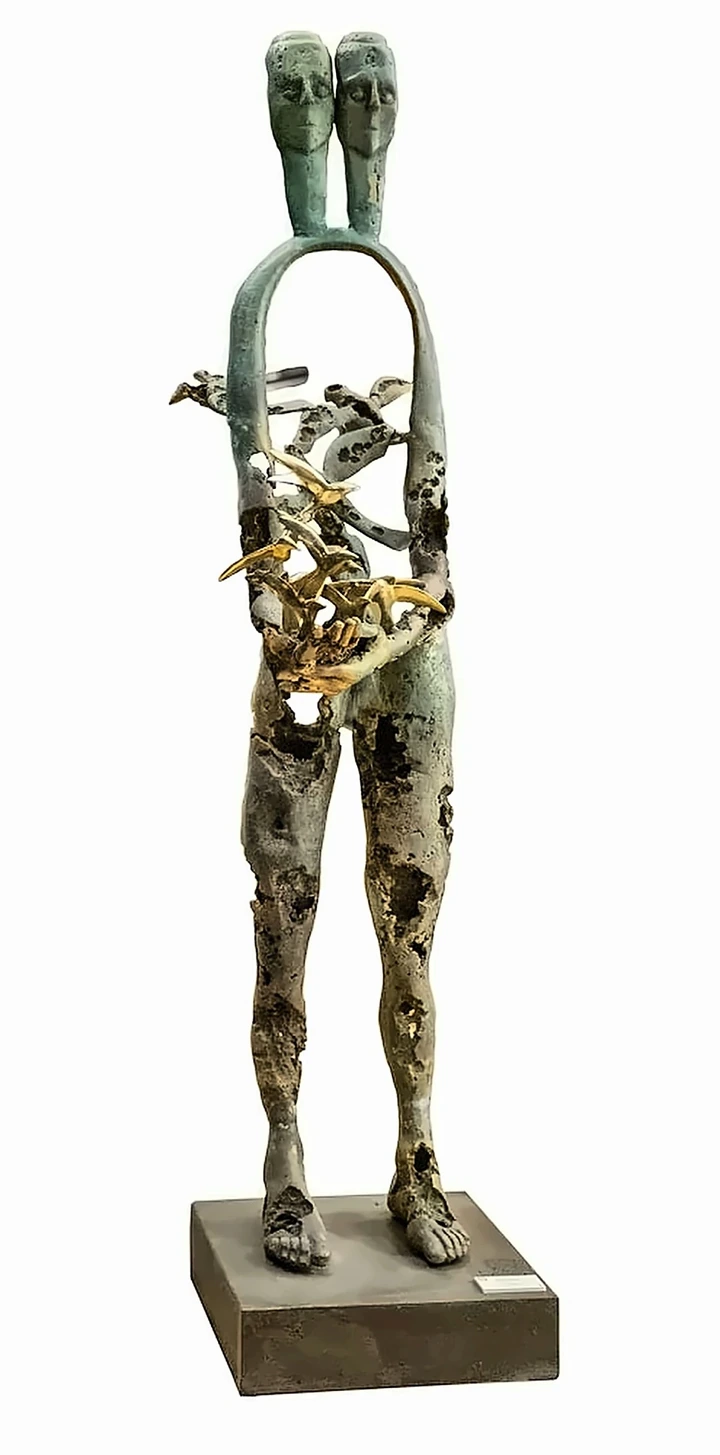 A tall bronze figure with two heads on one neck, its torso an open arch, holding a mass of gilded birds in both hands, the metal pitted and holed throughout.