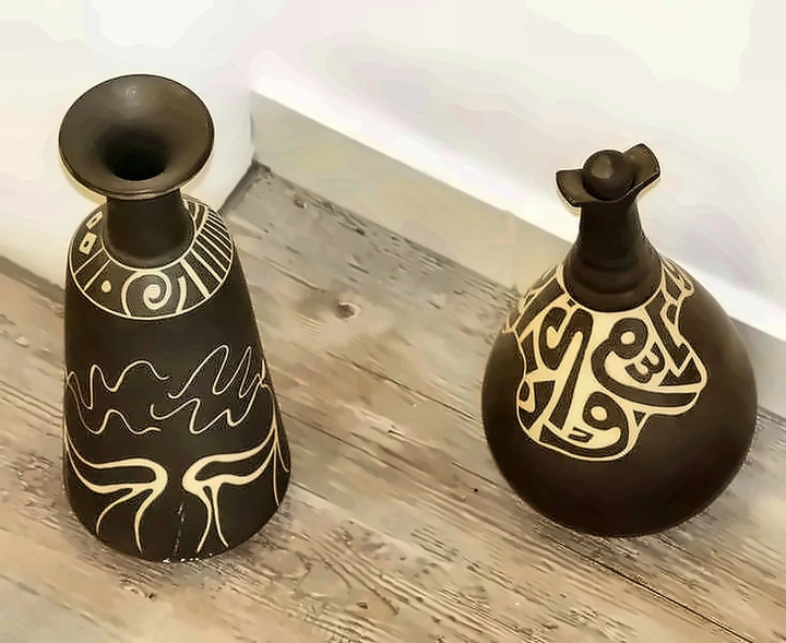 Two dark vessels, one conical and one round, cut through with white sgraffito line and script.