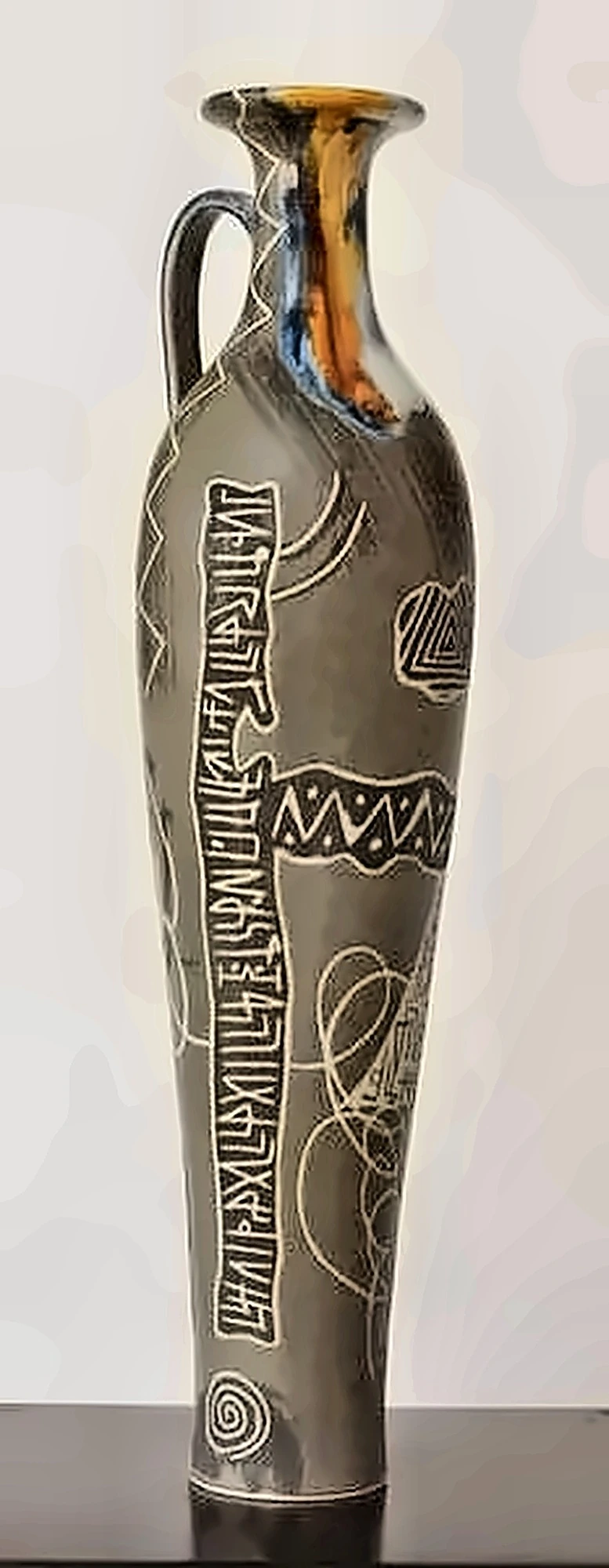 A tall grey jug with a handle, incised with vertical lines of script, colour running at the neck.