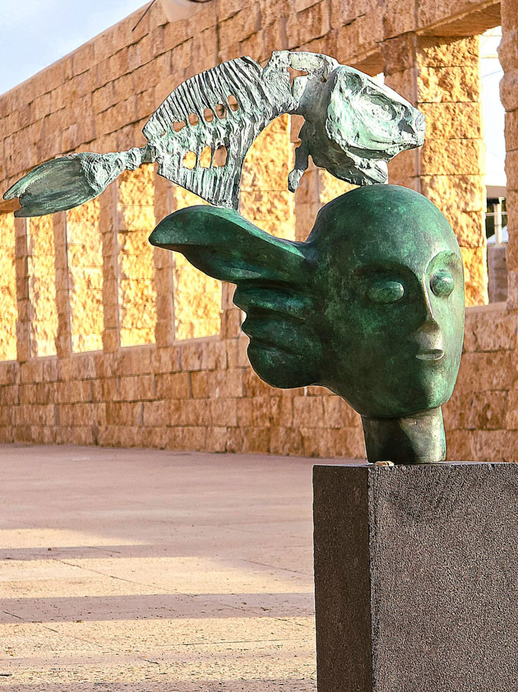 A sculpted head topped by a fish, outdoors; second angle.