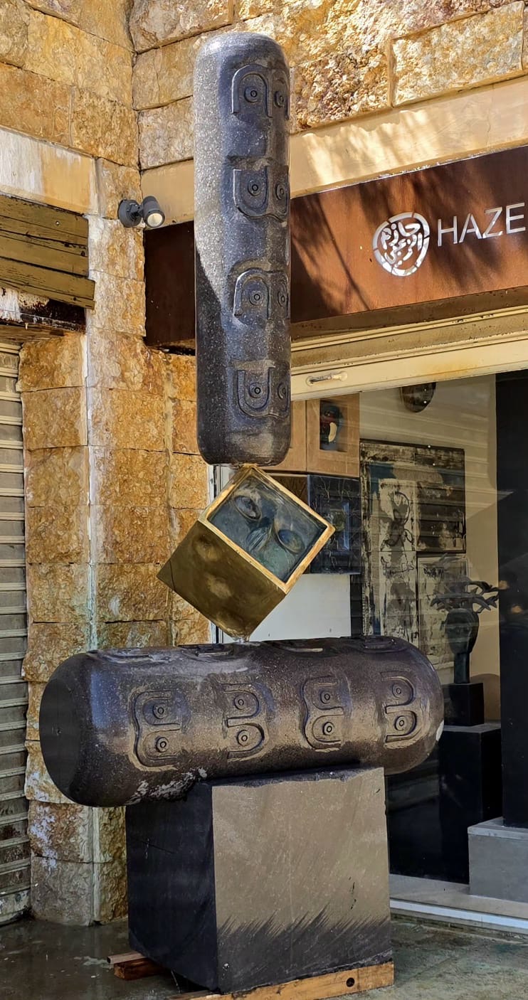 Basalt-and-bronze balancing sculpture outside the gallery, with a golden cube between two carved cylinders.