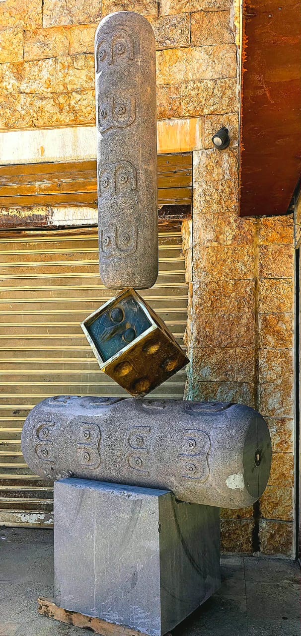 Side view of the balancing sculpture: carved basalt cylinders and a tilted bronze cube on a dark plinth.