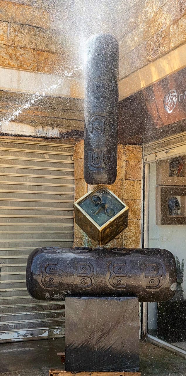 Water sprays across the upright basalt cylinder and golden cube, with the gallery entrance behind.