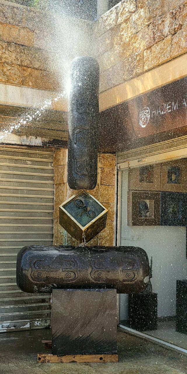 Wide view of the basalt-and-bronze sculpture under a shower of water in front of the gallery windows.