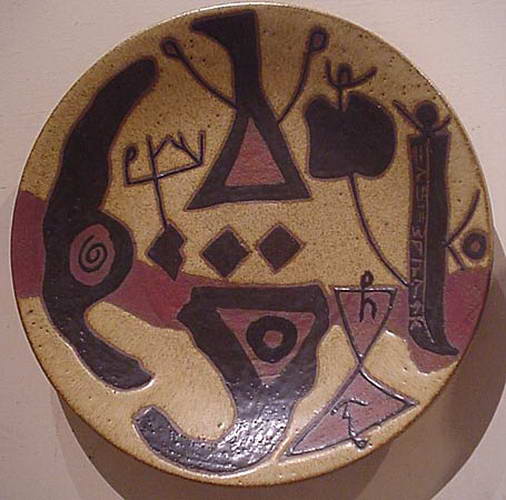 A stoneware plate with heavy black forms, a triangle, a spiral and a column of incised script down one side.