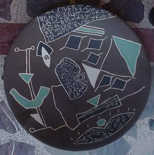 A dark grey plate with pale incised diamonds, blades and a band of script across the top, touched with turquoise.